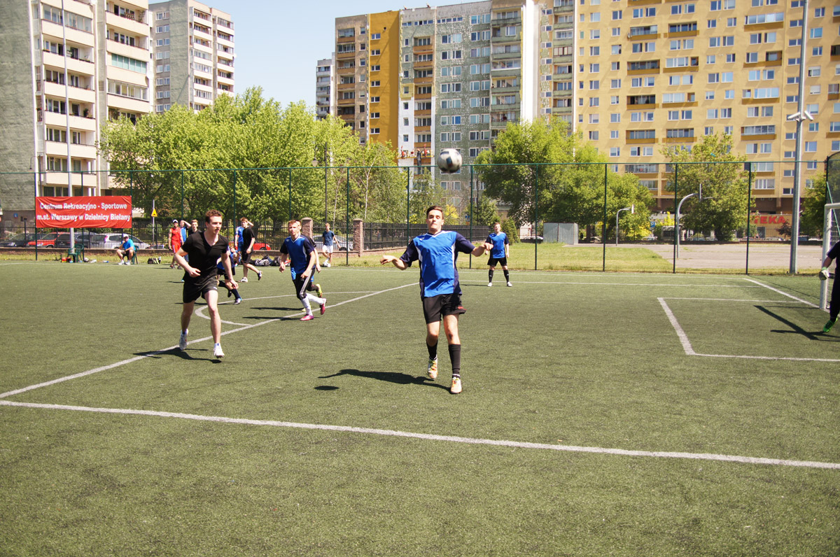 Kilku młodych chłopców grających w piłkę nożną na zielonej murawie ogrodzonego, zewnętrznego obiektu sportowego. W tle budynek gospodarczy.
