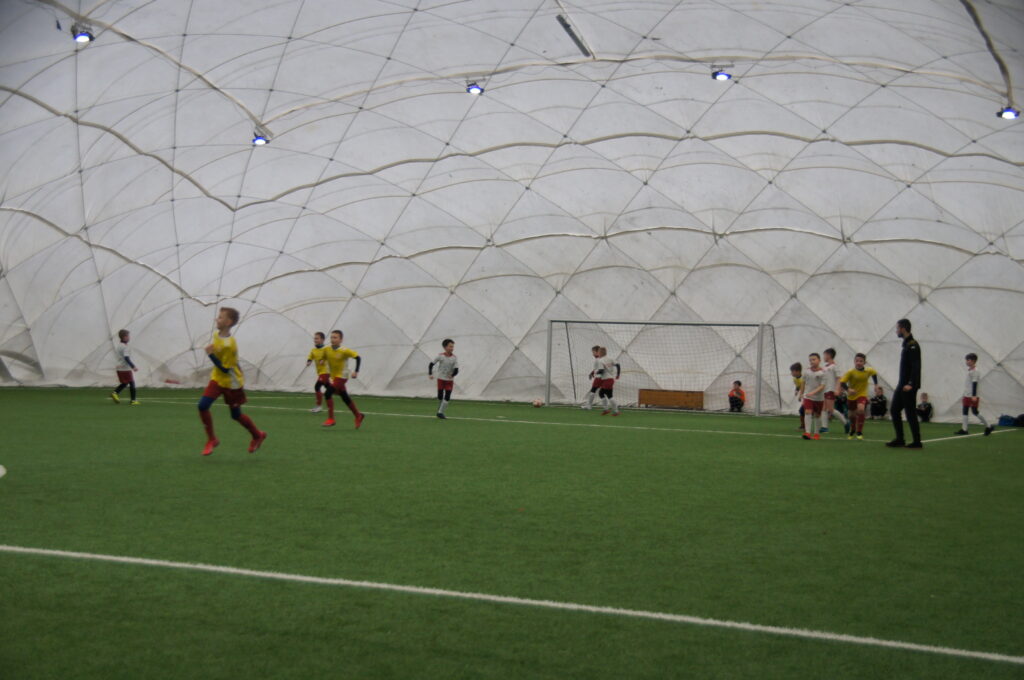 Kilkoro dzieci grających w piłkę nożną pod zadaszoną halą pneumatyczną w kolorze białym na zielonej murawie.