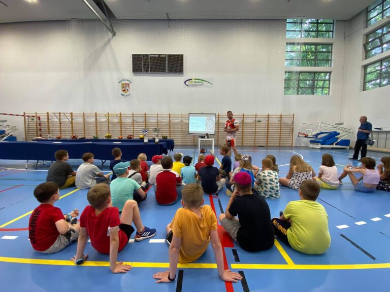Grupa dzieci słuchających pogadanki na hali sportowej.