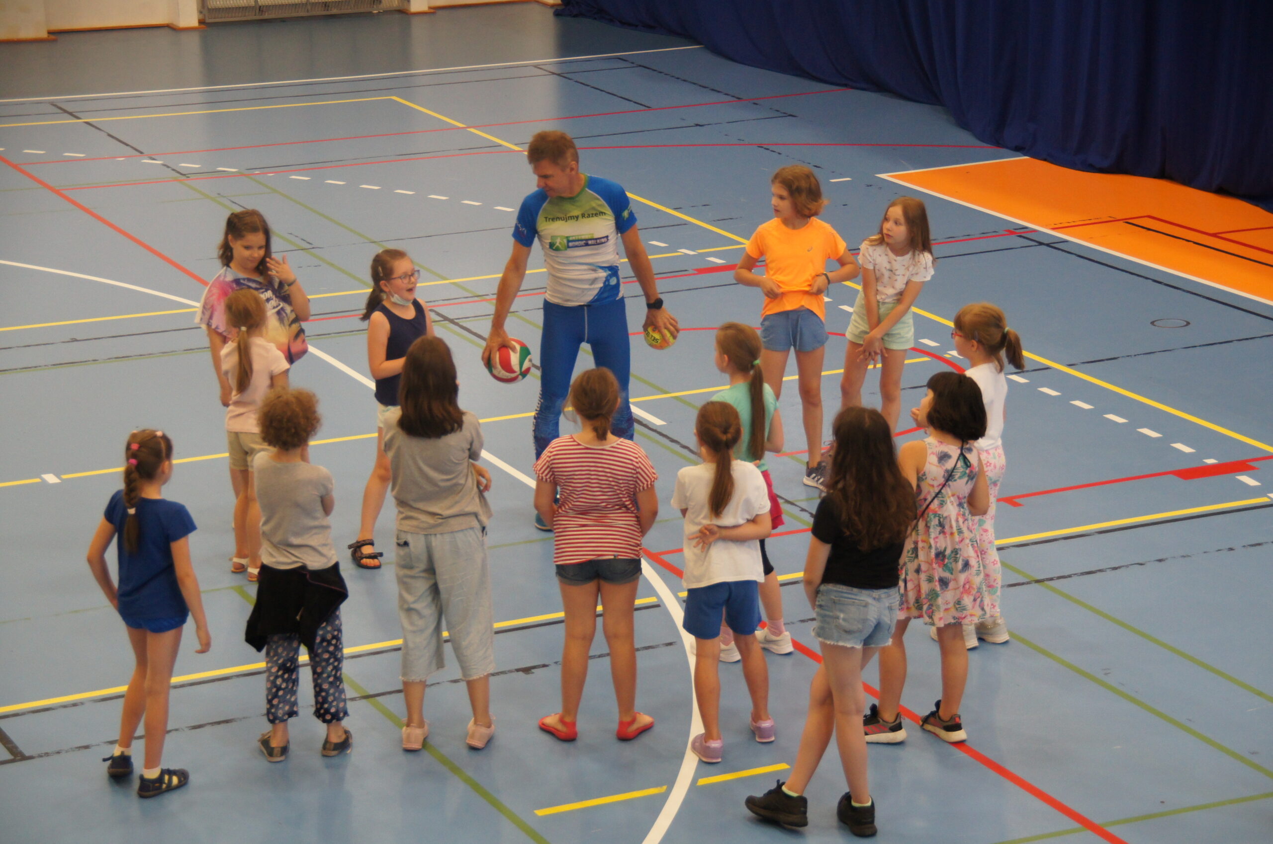 Grupa dzieci trenująca i bawiąca się na hali sportowej wraz z trenerem.