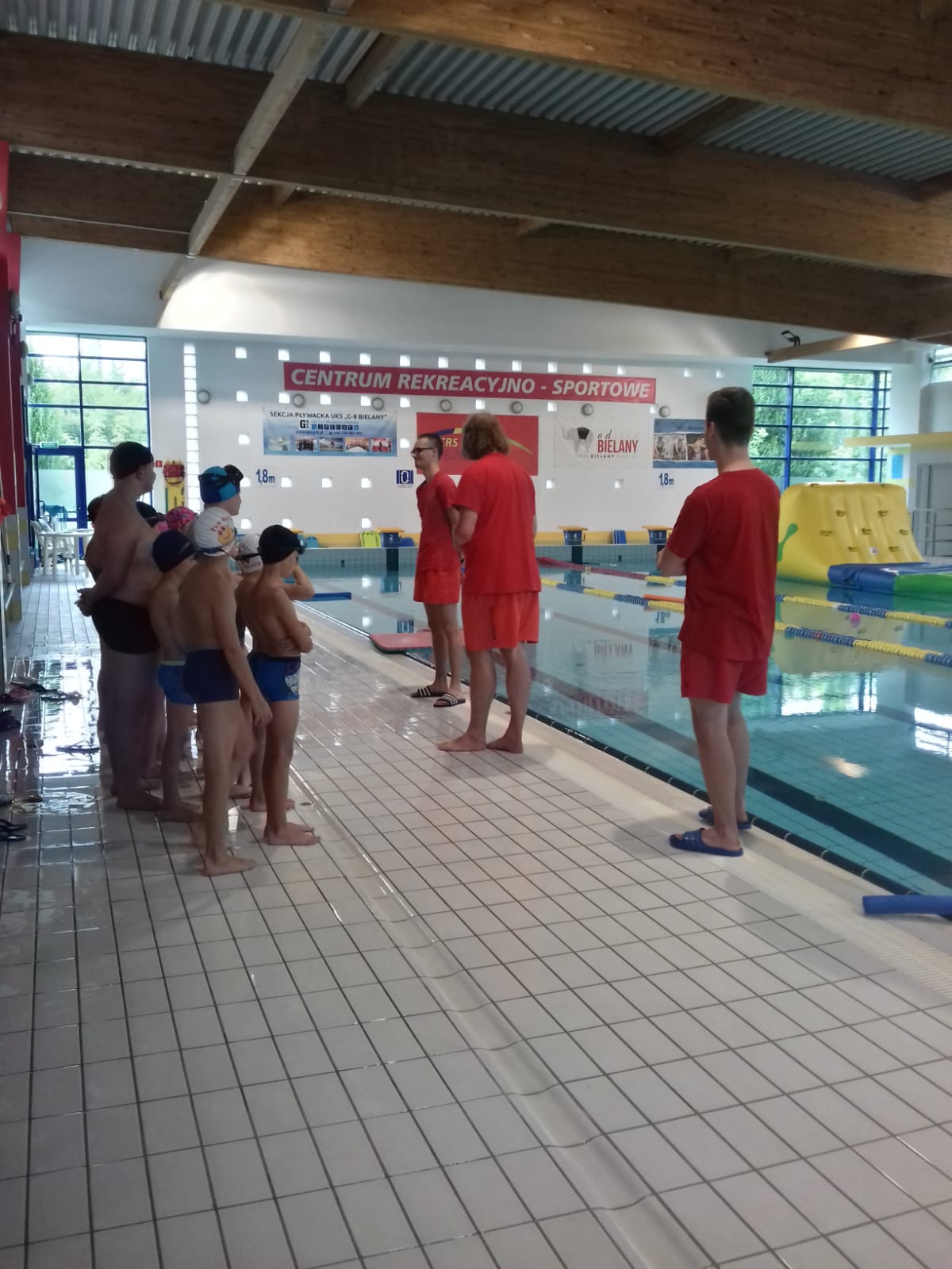 Grupa dzieci na pływalni słucha pogadanki na temat bezpieczeństwa.