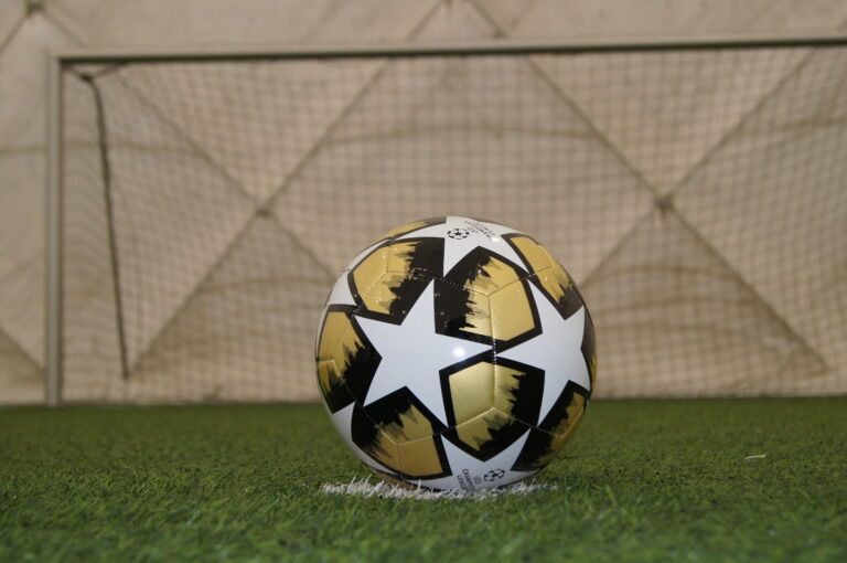 Piłka do piłki nożnej z kolorystyce biało, złoto, czarnej na tle bramki.