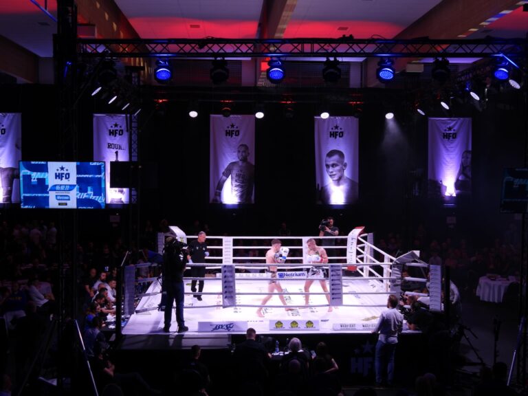 podświetlony ring z zawodnikami podczas gali, w tle zdjęcia zawodników na podświetlanych banerach.